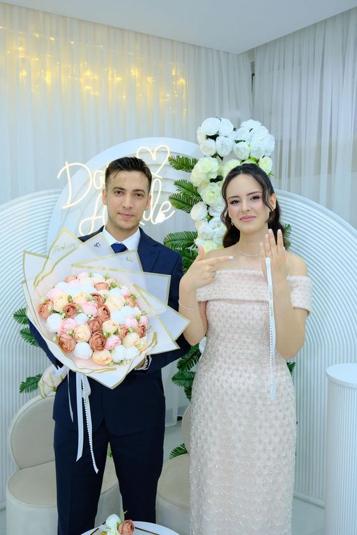 Çiçek buketi ve yüzüğüyle fotoğraf köşesinde poz veren nişanlı çift.
