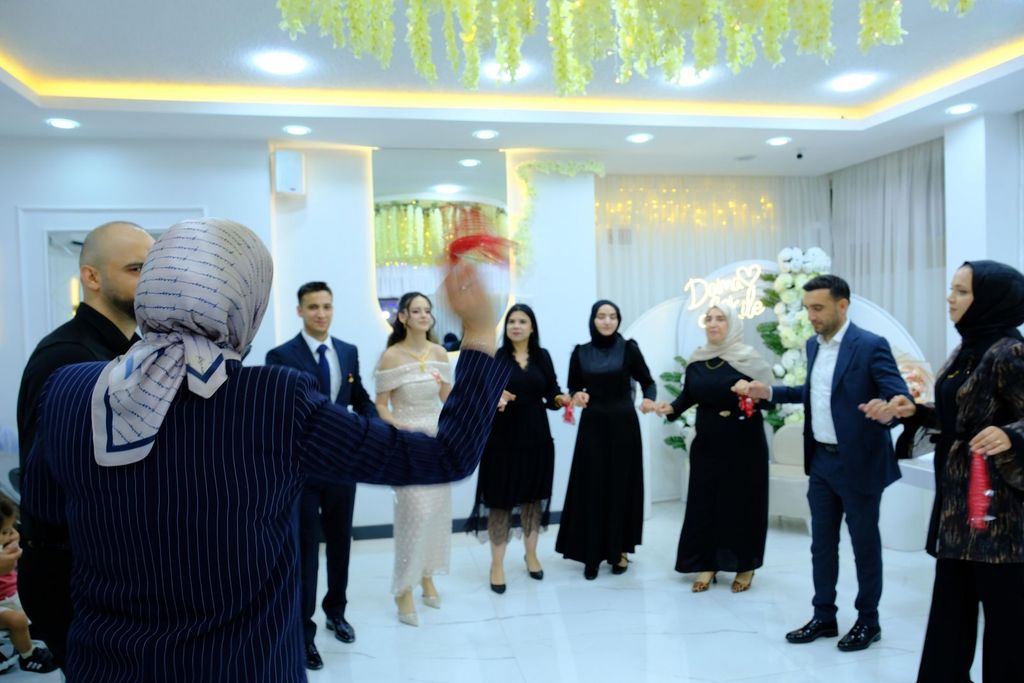Kapalı salonda el ele tutuşarak halay çeken nişan davetlileri ve çift.