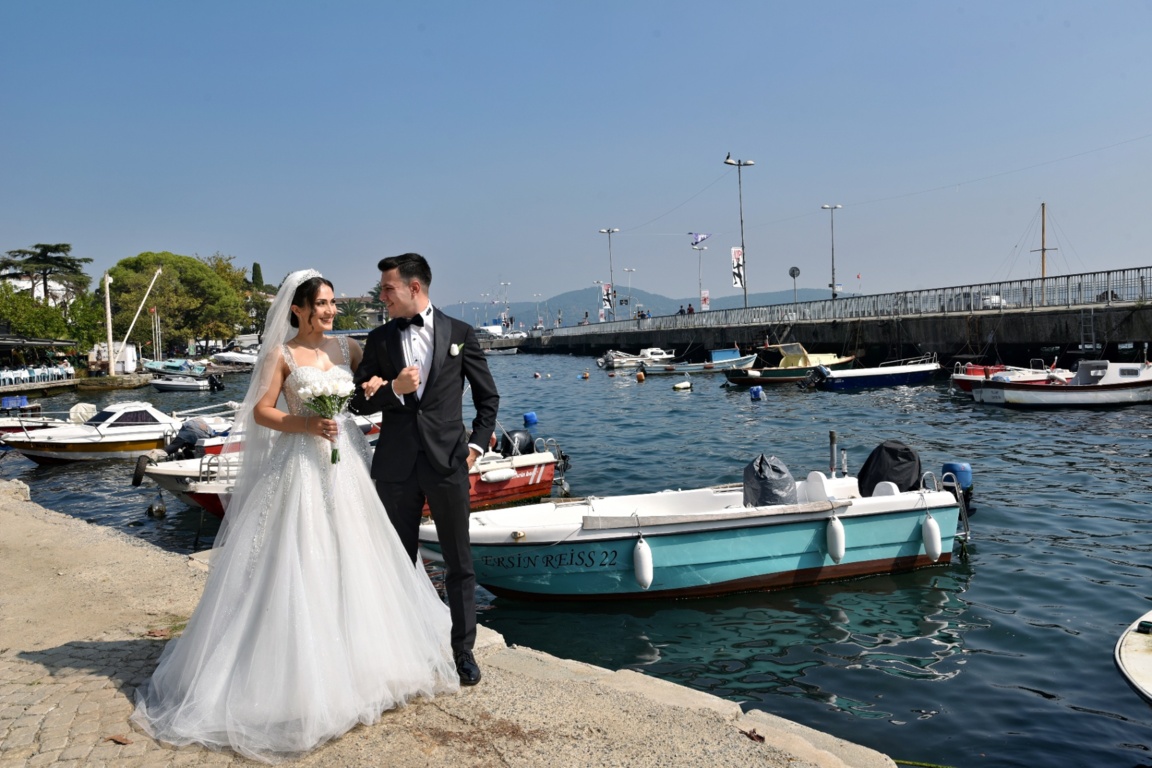 Deniz kenarında teknelerin önünde kol kola duran gelin ve damat.