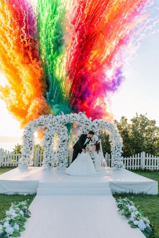 Çiçekli nikah takı önünde renkli duman gösterisi eşliğinde duran gelin ve damat.